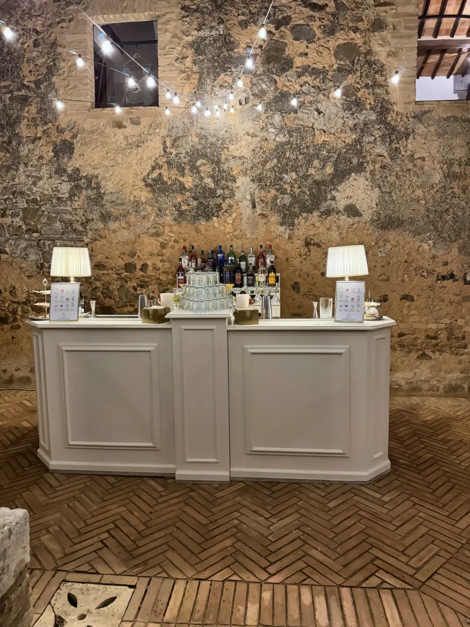 White bar with lamps under string lights, stone courtyard in Montalcino