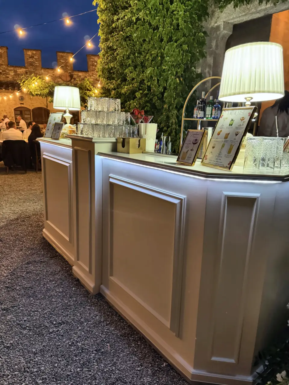 Evening bar under string lights, wedding dinner in the background